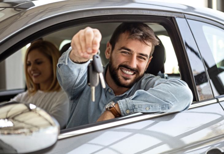 A smiling man in a light denim shirt leans out the driver’s side window of a new car, proudly holding up a set of keys. A woman sits beside him in the passenger seat, also smiling. This confident moment showcases the satisfaction of buying a vehicle from The Auto Professionals of Indianapolis, Indiana. Visit theautoprofessionals.com to learn more!