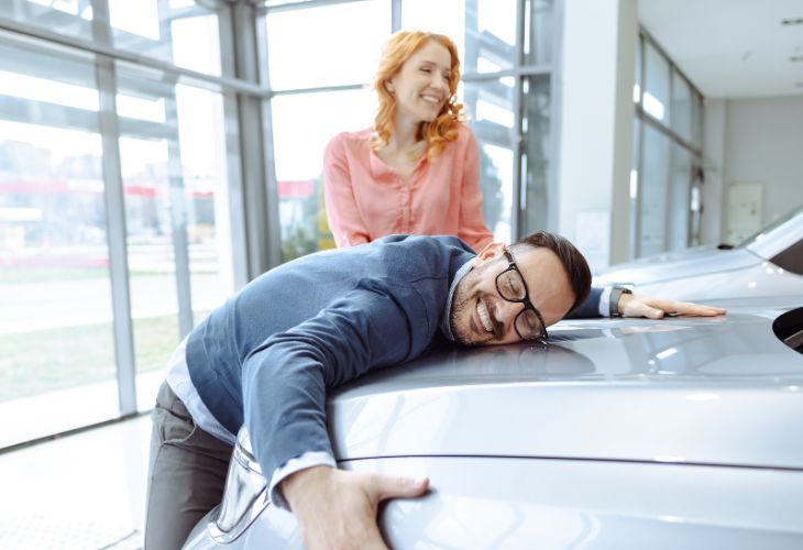 A cheerful man in glasses and a blue blazer leans over and hugs the hood of a shiny silver car with a huge smile on his face. A red-haired woman in a pink blouse stands behind him, laughing with joy. Their playful excitement reflects the happiness of finding the perfect vehicle with The Auto Professionals of Indianapolis, Indiana. Visit theautoprofessionals.com to learn more!