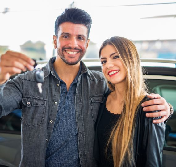 A happy couple stands in front of a shiny red SUV inside a bright car dealership. The man, dressed in a light blue button-up and jeans, holds up a car key with pride, while the woman, in a white blouse and beige pants, wraps her arms around him with a joyful smile. Their excitement captures the satisfaction of buying a vehicle from a trusted team like The Auto Professionals of Indianapolis, Indiana. Visit theautoprofessionals.com to learn more!